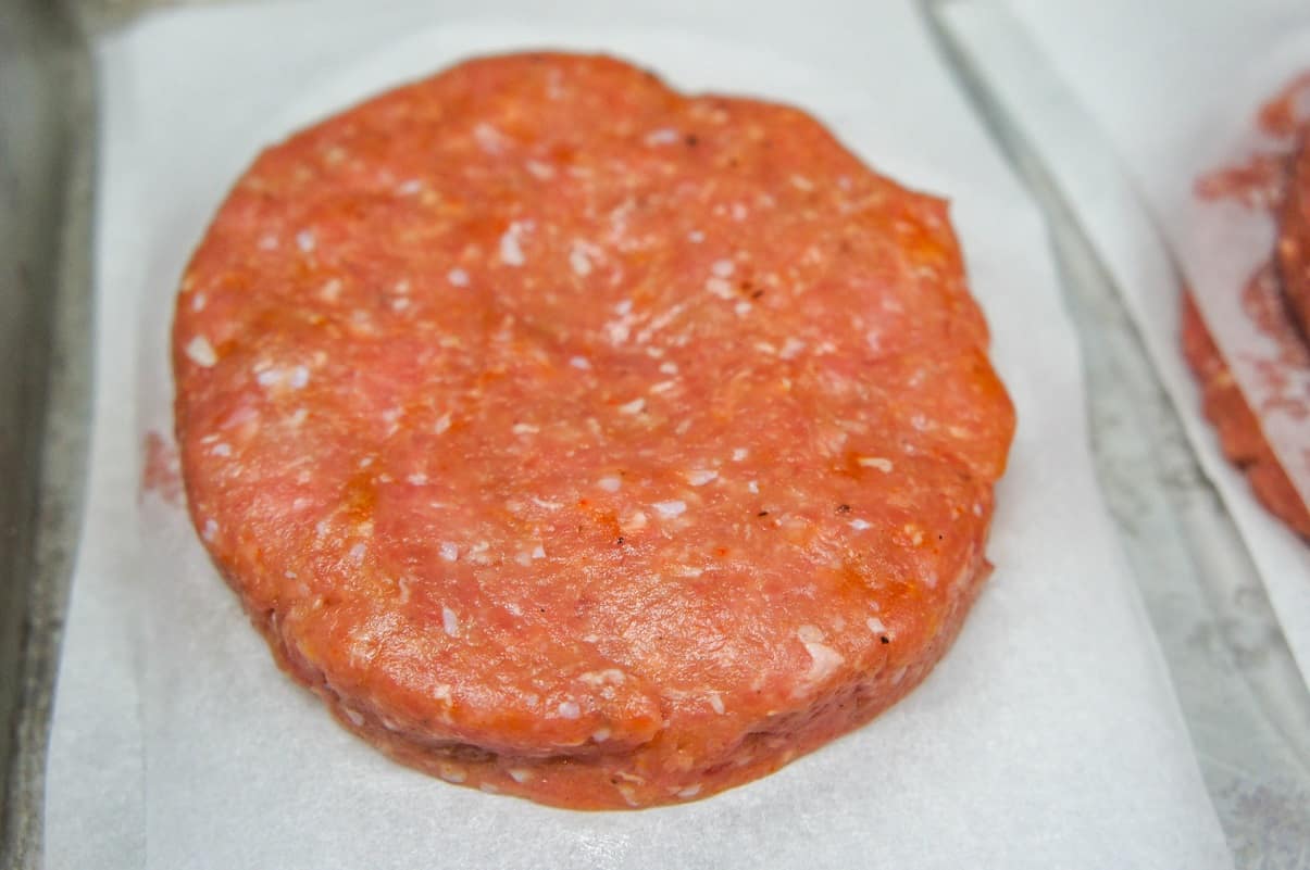 A formed burger patty, before cooking on a square piece of white parchment paper.