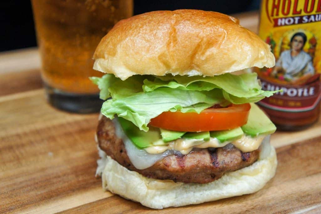 A burger stack high with avocado, tomato and lettuce on a wood cutting board with a beer glass and hot sauce in the background.