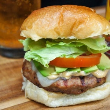 A burger stack high with avocado, tomato and lettuce on a wood cutting board with a beer glass and hot sauce in the background.