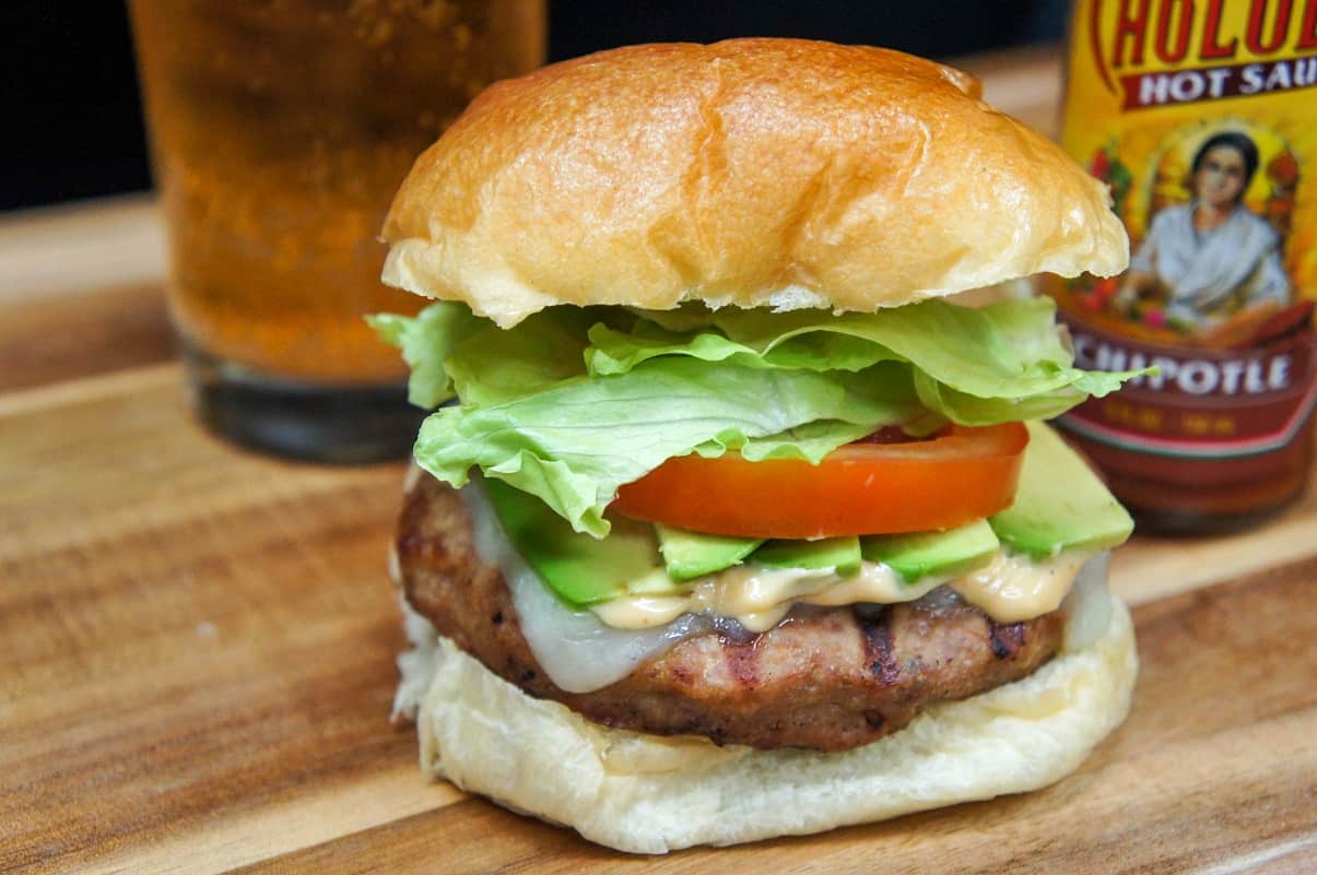 A burger stack high with avocado, tomato and lettuce on a wood cutting board with a beer glass and hot sauce in the background.