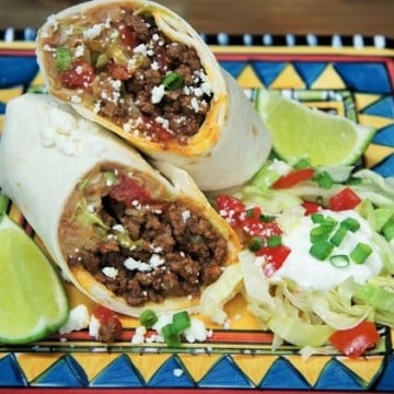 Beef & Bean Burritos, cut in half and served with a side of shredded lettuce, tomatoes, green onions and lime wedges on a colorful plate