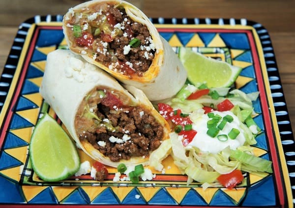Beef & Bean Burritos, cut in half and served with a side of shredded lettuce, tomatoes, green onions and lime wedges on a colorful plate