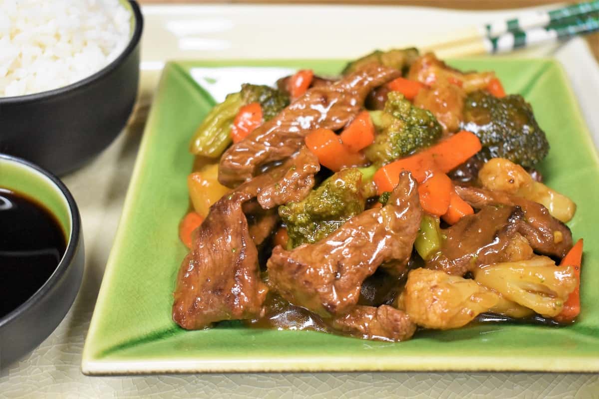 Beef and vegetable stir fry served on a green plate with a side of white rice and soy sauce.