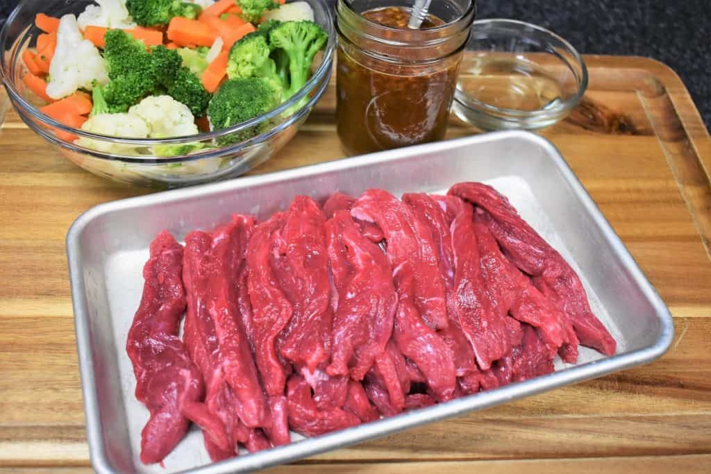 Sliced beef, a bowl of broccoli, cauliflower and carrots and stir fry sauce displayed on a wood cutting board.