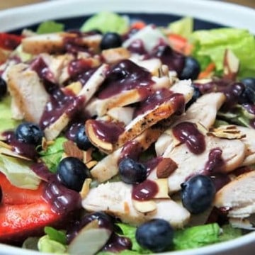 Berry Chicken Salad, a bed of lettuce topped with sliced strawberries, blueberries, thin sliced chicken and drizzed with a blueberry dressing, served in a blue bowl with a white rim