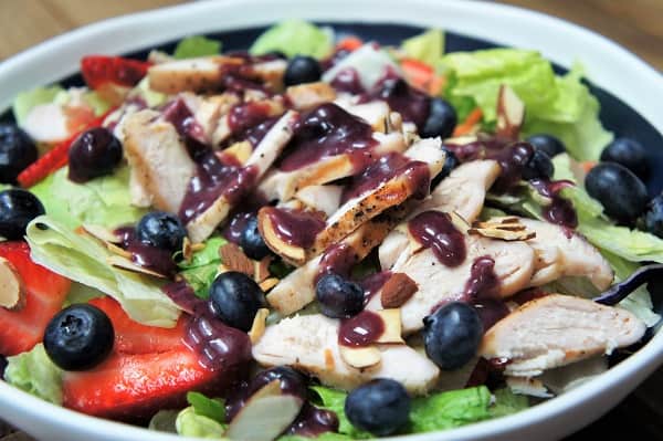 Berry Chicken Salad, a bed of lettuce topped with sliced strawberries, blueberries, thin sliced chicken and drizzed with a blueberry dressing, served in a blue bowl with a white rim