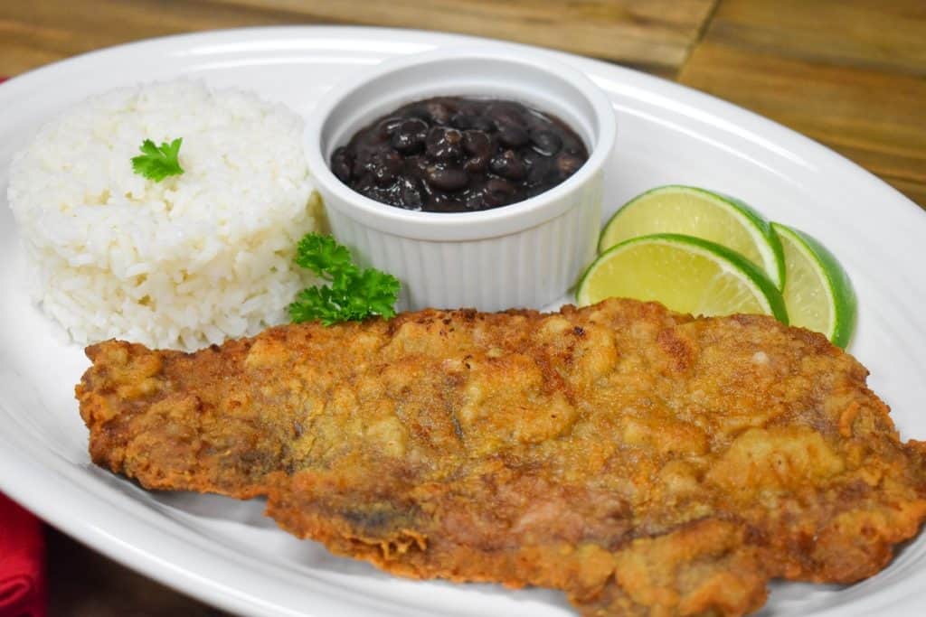 Bistec empanizado served on a large white platter with a side of white rice, black beans and lime wedges.