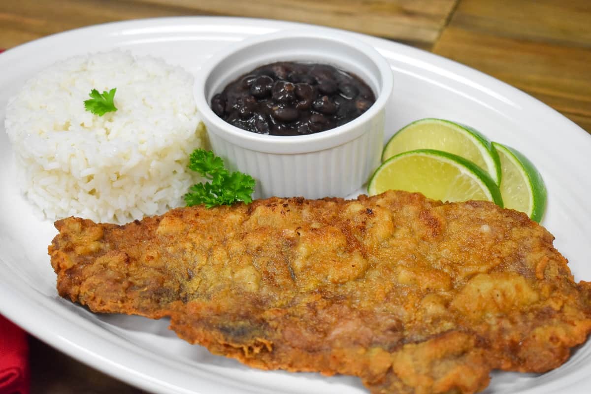 Bistec empanizado served on a large white platter with a side of white rice, black beans and lime wedges.