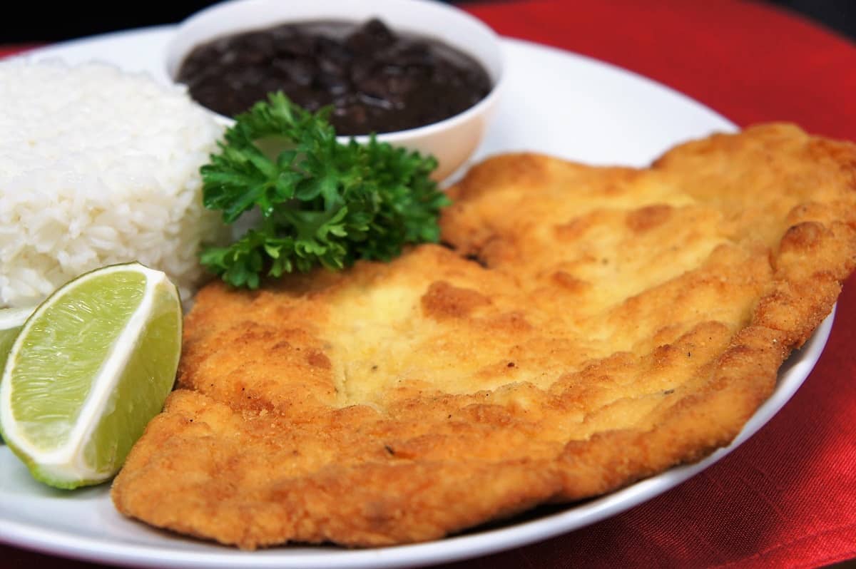 Breaded chicken steak, a thin chicken cutlet that is breaded and fried, served with white rice and black beans on a white plate.