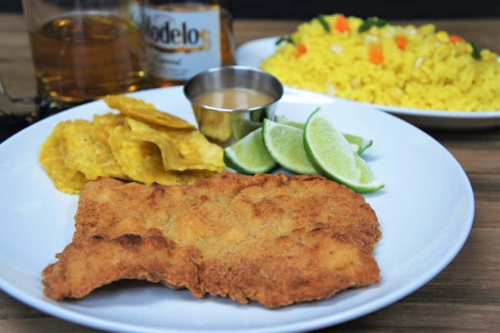 Breaded Pork Steak on a white plate served with plantain chips and lime wedges. A half-filled glass and bottle of beer and yellow rice are in the background.