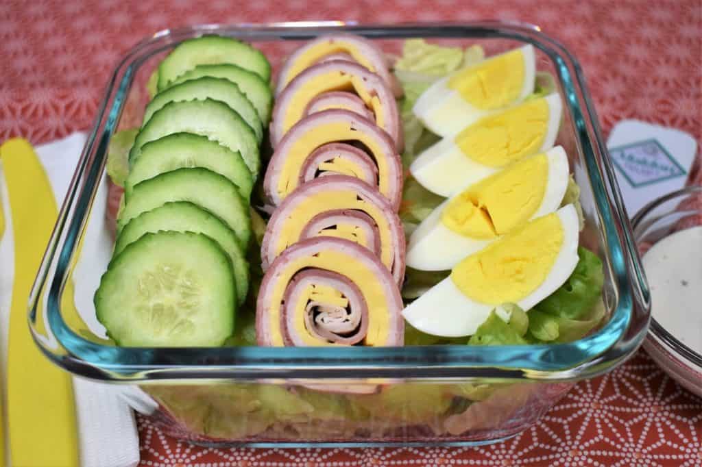 Chef salad with sliced cucumbers, ham, turkey and cheese roll-ups and a quartered hard boiled egg all on top of a bed of lettuce. The salad is in a clear glass, square container.