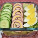 Chef salad with sliced cucumbers, ham, turkey and cheese roll-ups and a quartered hard boiled egg all on top of a bed of lettuce. The salad is in a clear glass, square container.