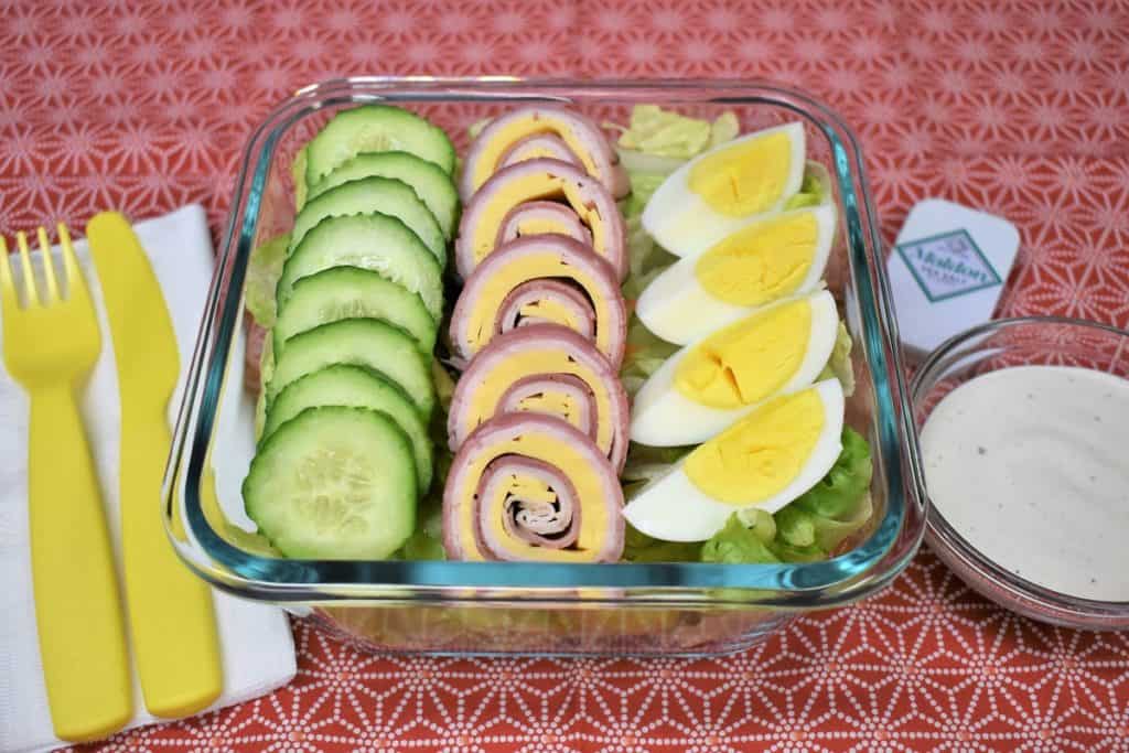 Chef salad with sliced cucumbers, ham, turkey and cheese roll-ups and a quartered hard boiled egg all on top of a bed of lettuce. The salad is in a clear glass, square container with yellow plastic utensils and ranch dressing on the side.