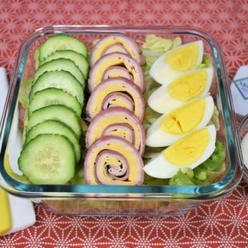 Chef salad with sliced cucumbers, ham, turkey and cheese roll-ups and a quartered hard boiled egg all on top of a bed of lettuce. The salad is in a clear glass, square container with yellow plastic utensils and ranch dressing on the side.