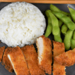 Chicken Katsu thin chicken breast breaded with panko and fried and sliced into strips served on a gray plate with a side of white rice and edamame.