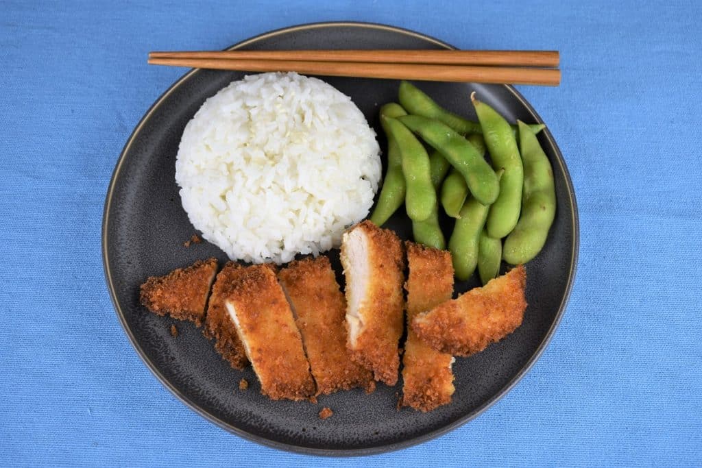 Chicken Katsu thin chicken breast breaded with panko and fried and sliced into strips served on a gray plate with a side of white rice and edamame.