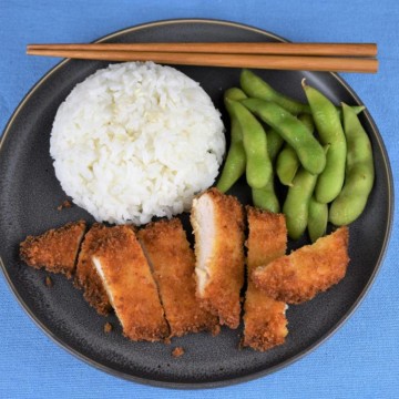 Chicken Katsu thin chicken breast breaded with panko and fried and sliced into strips served on a gray plate with a side of white rice and edamame.