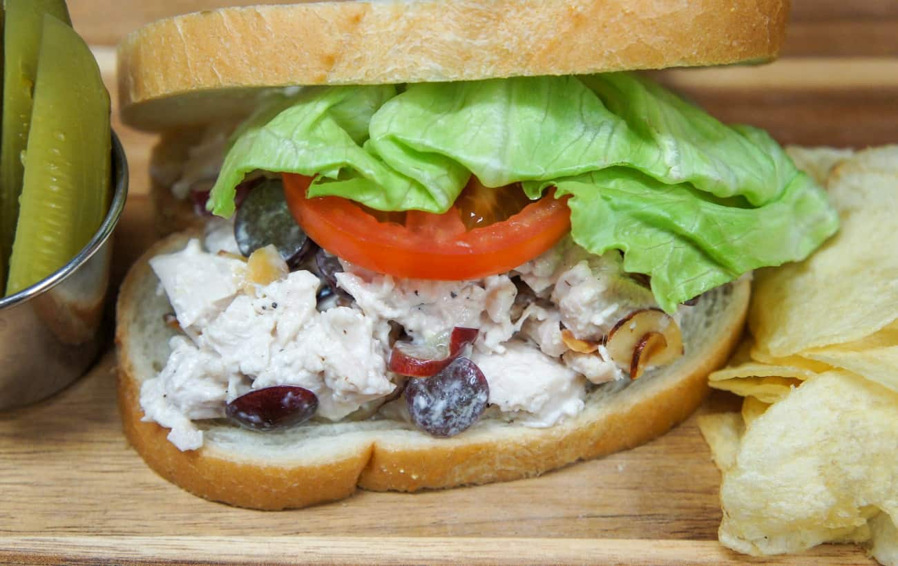 A close up image of the chicken salad on white bread, lettuce and tomato with pickle spears and potato chips on the side.