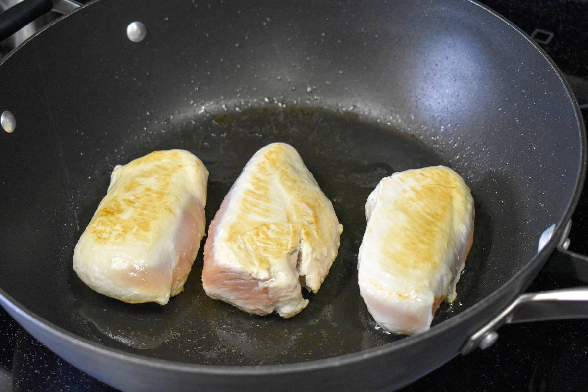 Three chicken breasts browning in a large, black skillet.