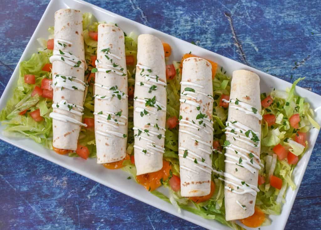 Five taquitos arranged on a white platter on a bed of shredded lettuce with diced tomatoes. They are garnished with a little sour cream and chopped parsley.