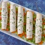 Five taquitos arranged on a white platter on a bed of shredded lettuce with diced tomatoes. They are garnished with a little sour cream and chopped parsley.