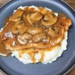 Thin chicken breasts covered in brown mushroom gravy served on a bed of mashed potatoes on a gray plate.
