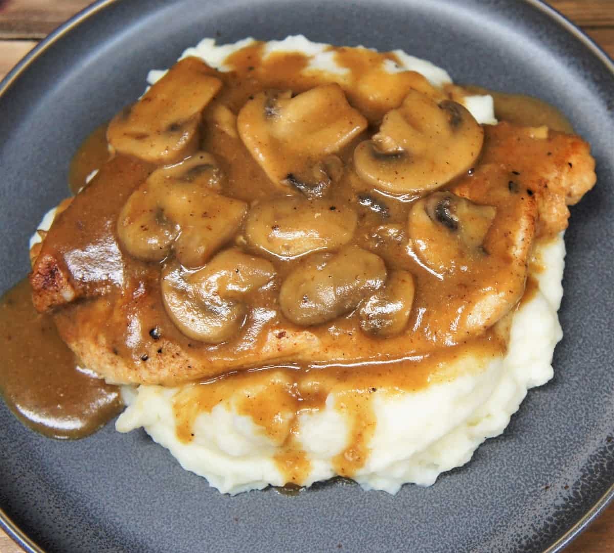 Thin chicken breasts covered in brown mushroom gravy served on a bed of mashed potatoes on a gray plate.