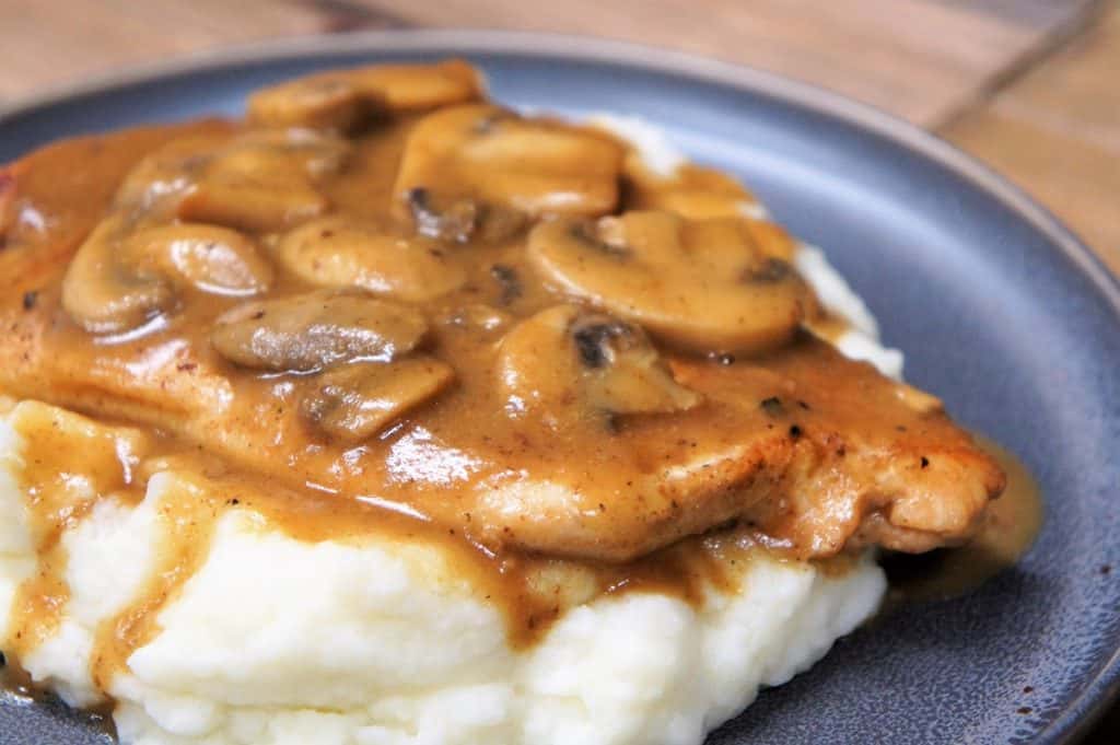 Thin chicken breasts covered in brown mushroom gravy served on a bed of mashed potatoes on a gray plate.