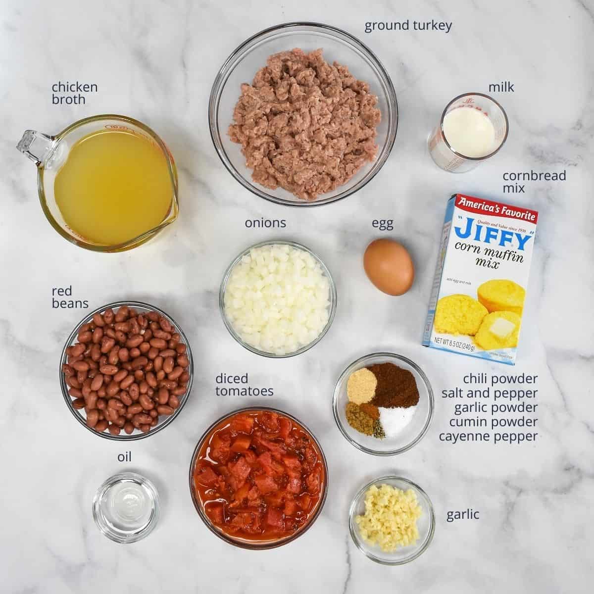 The prepped ingredients for the casserole in glass bowls (except the cornbread mix is in its box) arranged on a white table. Each ingredient is labeled on the top or side with the name.