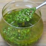 Chimichurri sauce in a clear glass container with a small spoon.