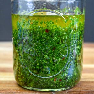 Chimichurri in a glass mason jar set on a wood cutting board.