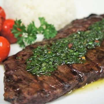 Churrasco & Chimichurri, steak topped with chimichurri sauce served on a white plate with a side of grilled tomatoes and white rice