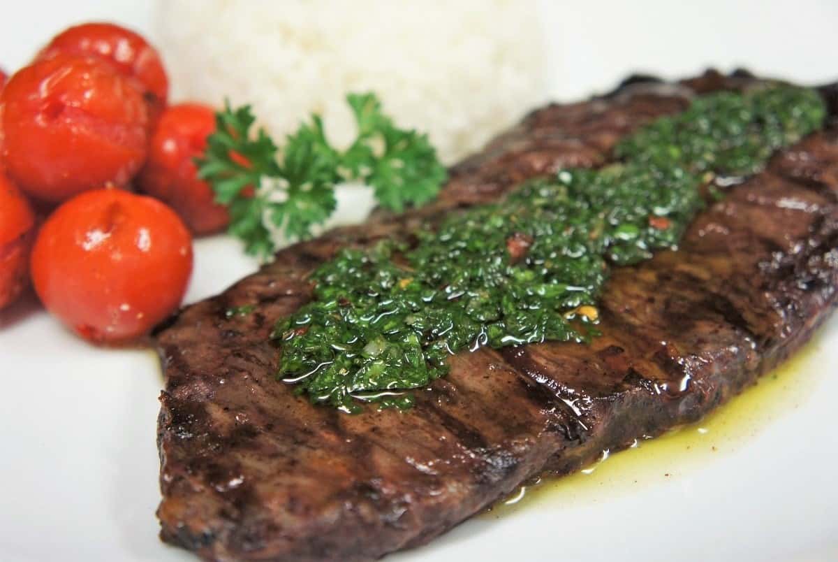 Churrasco & Chimichurri, steak topped with chimichurri sauce served on a white plate with a side of grilled tomatoes and white rice