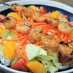 A bed of lettuce topped with tomato wedges, mandarin orange slices, shredded carrots and two grilled shrimp skewers, served in a blue bowl with a white rim