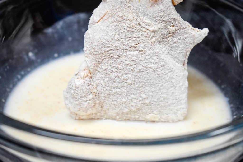 A chicken thigh that has been coated in flour being dunked into a bowl of milk.