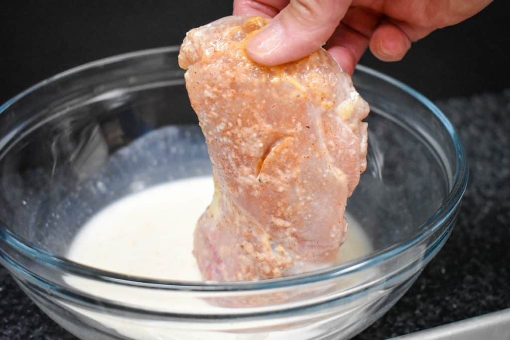 A moistened chicken thigh that had been coated in flour being pulled out of a bowl of milk.