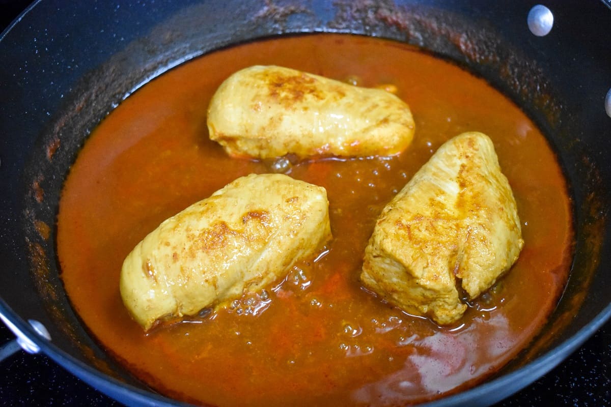 Three chicken breasts sitting in red sauce in a large, black skillet.