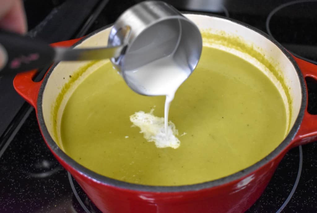 Half and half being added to the pureed asparagus soup. The soup is in a red cast iron pot.