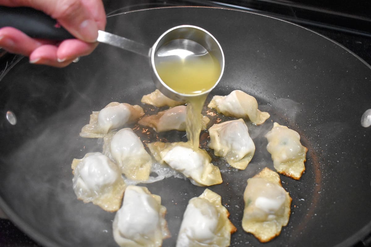 A quarter cup of chicken broth being added to potstickers in a large, black skillet.