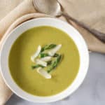 The cream of asparagus soup garnished with asparagus tips and parmesan cheese and served in a white bowl with a beige linen and a spoon.