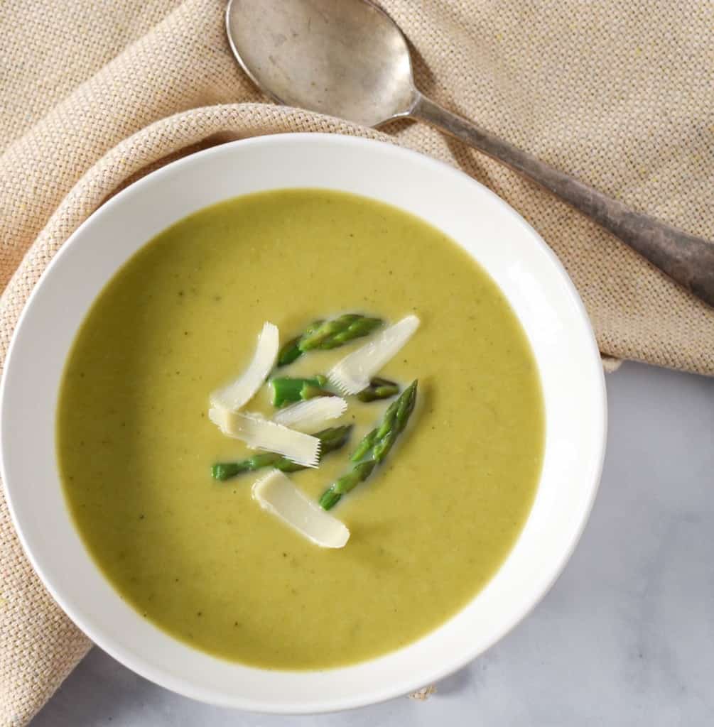 The cream of asparagus soup garnished with asparagus tips and parmesan cheese and served in a white bowl with a beige linen and a spoon.
