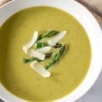 A close up image of the soup garnished with asparagus tips and parmesan cheese served in a white bowl.