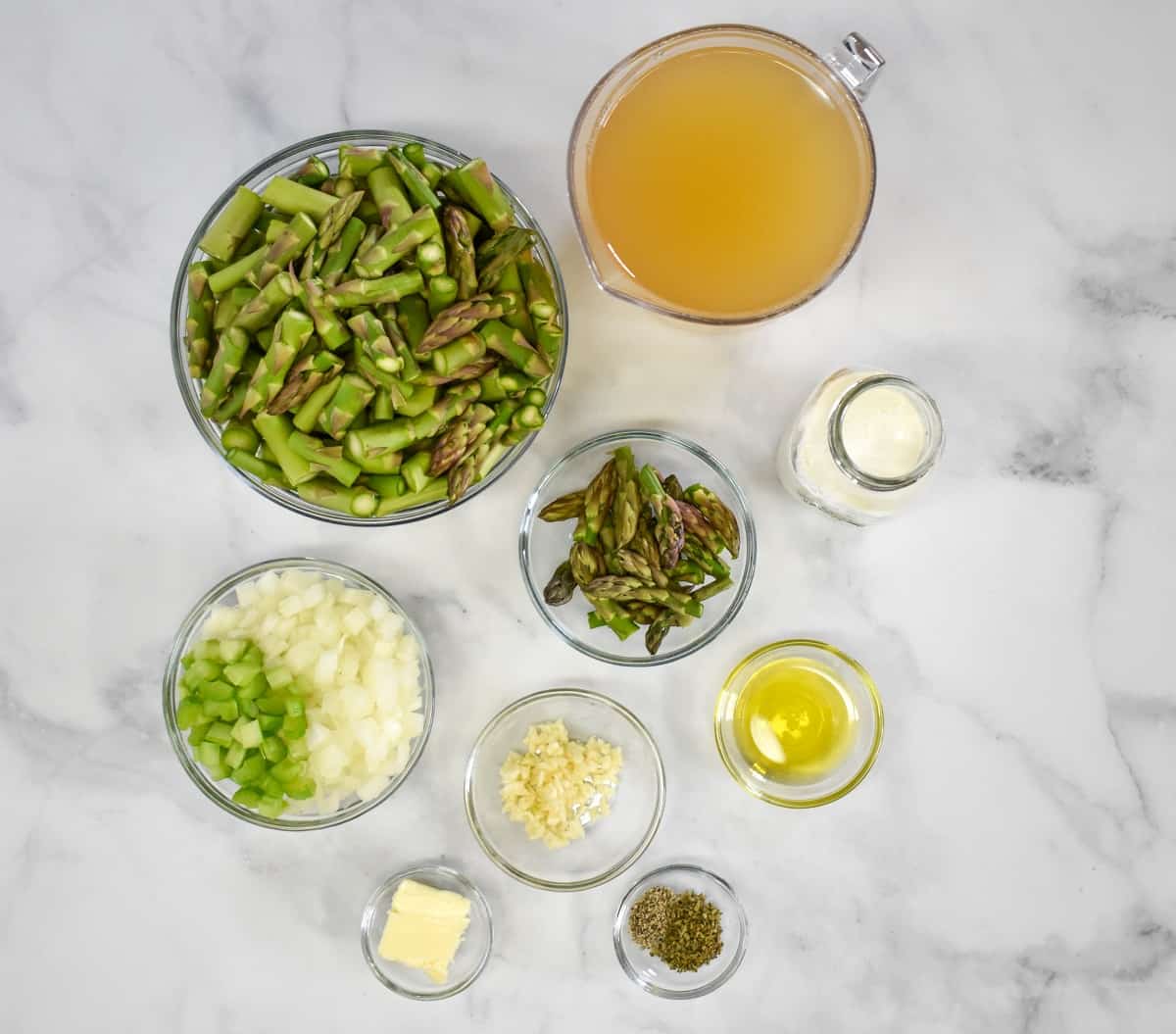 The ingredients for the cream of asparagus soup prepped and separated in glass bowls arranged on a white table.