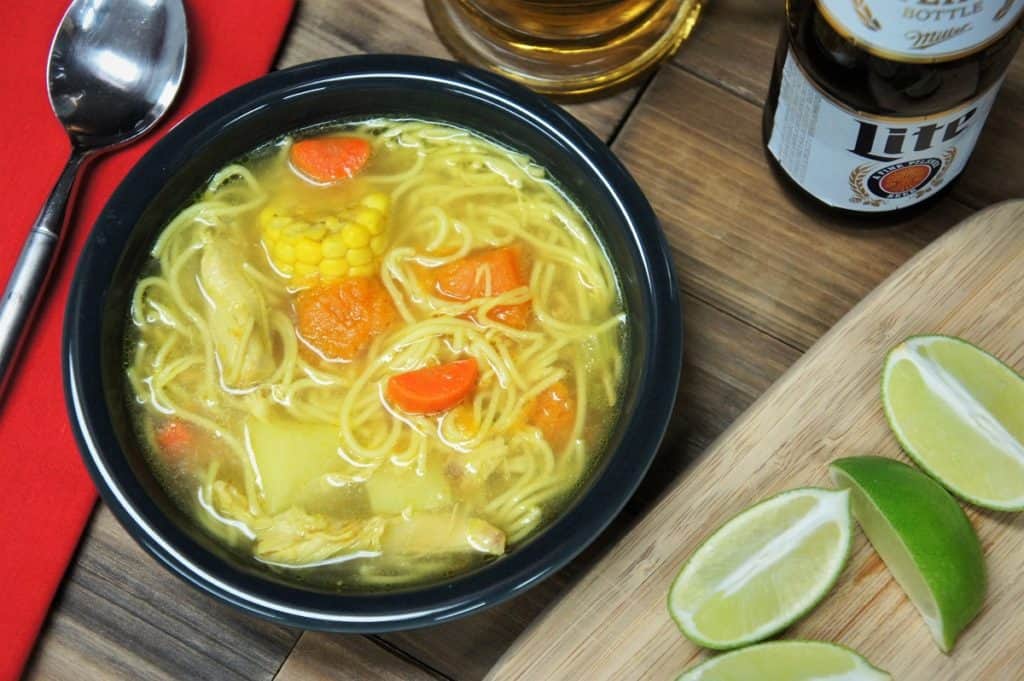 Cuban chicken soup with noodles carrots, potatoes, pumpkin, corn and chicken served in a dark gray bowl with lime wedges and a beer mug in the background.