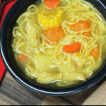 Cuban chicken soup with noodles carrots, potatoes, pumpkin, corn and chicken served in a dark gray bowl with lime wedges and a beer mug in the background.