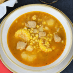 Cuban Corn Stew served in a white bowl with a bread basket and a beer mug in the background.