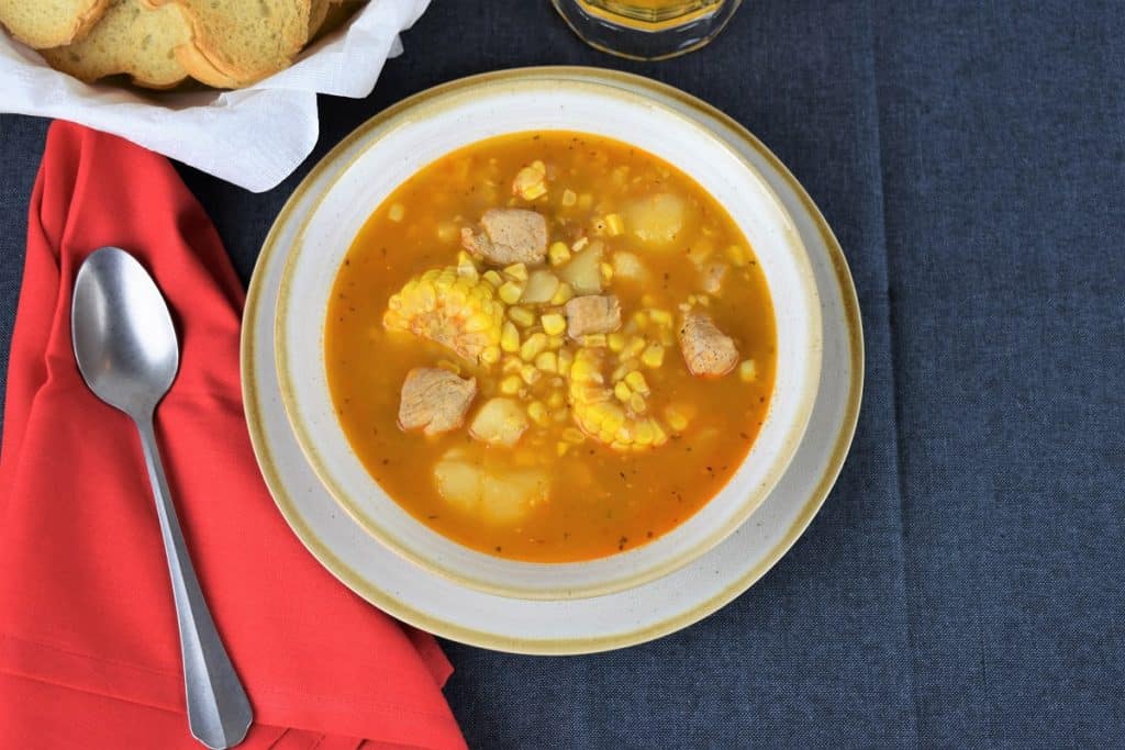 Cuban Corn Stew, or guiso de maiz in Spanish is served in a light colored bowl and loaded with corn, pork chunks, potatoes, served with a bread basket and beer in the background.