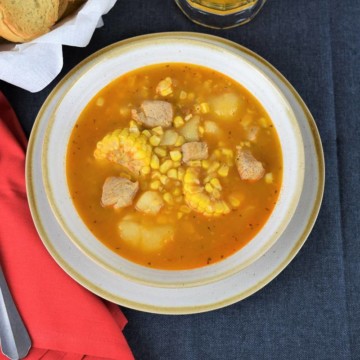 Cuban Corn Stew, or guiso de maiz in Spanish is served in a light colored bowl and loaded with corn, pork chunks, potatoes, served with a bread basket and beer in the background.