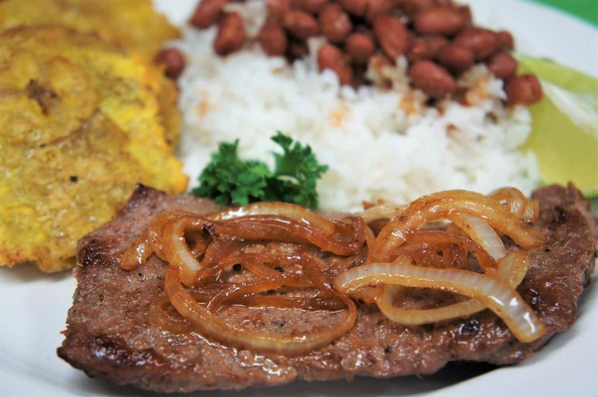 Cuban Cube Steak, topped with cooked onions and served with white rice, red beans and fried green plantains on the side.