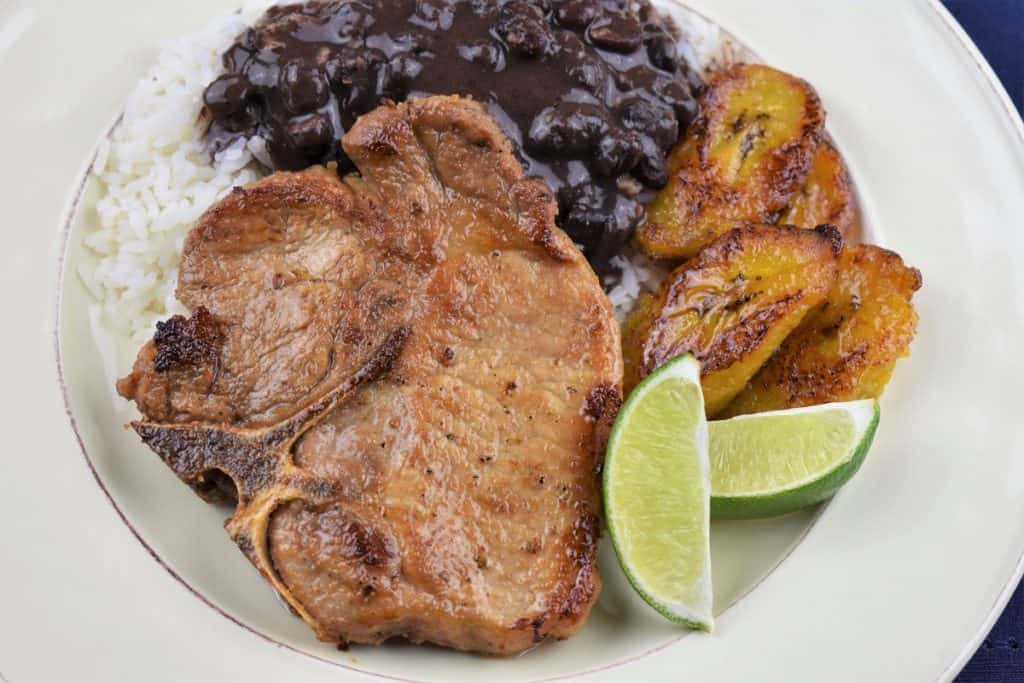 A thin cut, bone in pork chop served with white rice, black beans and fried sweet plantains on a white plate